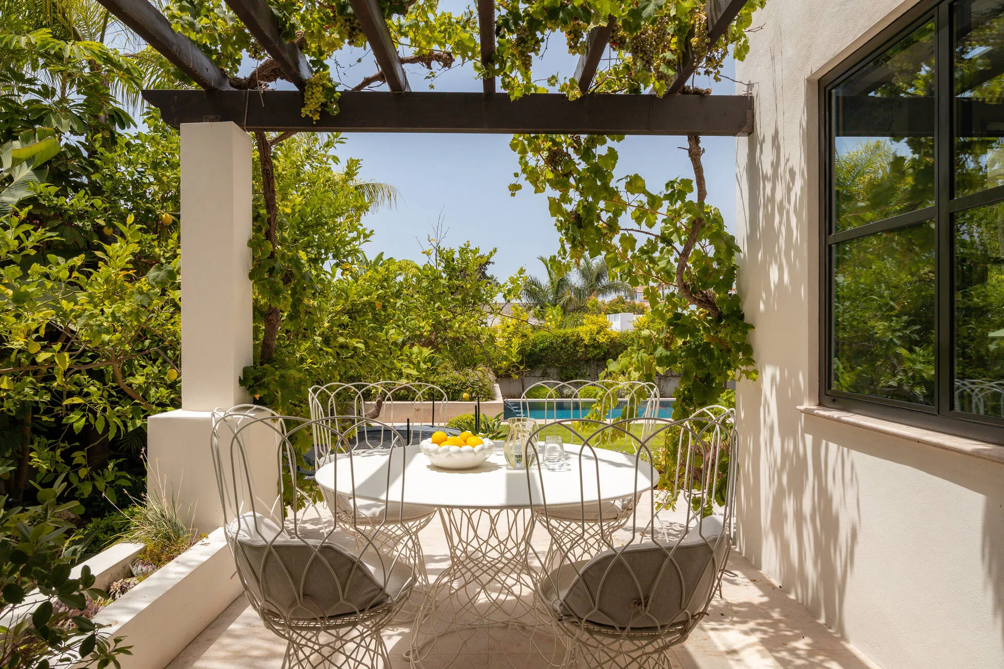 A round table beneath the grapevine pergola, lemons in a bowl, the pool beyond