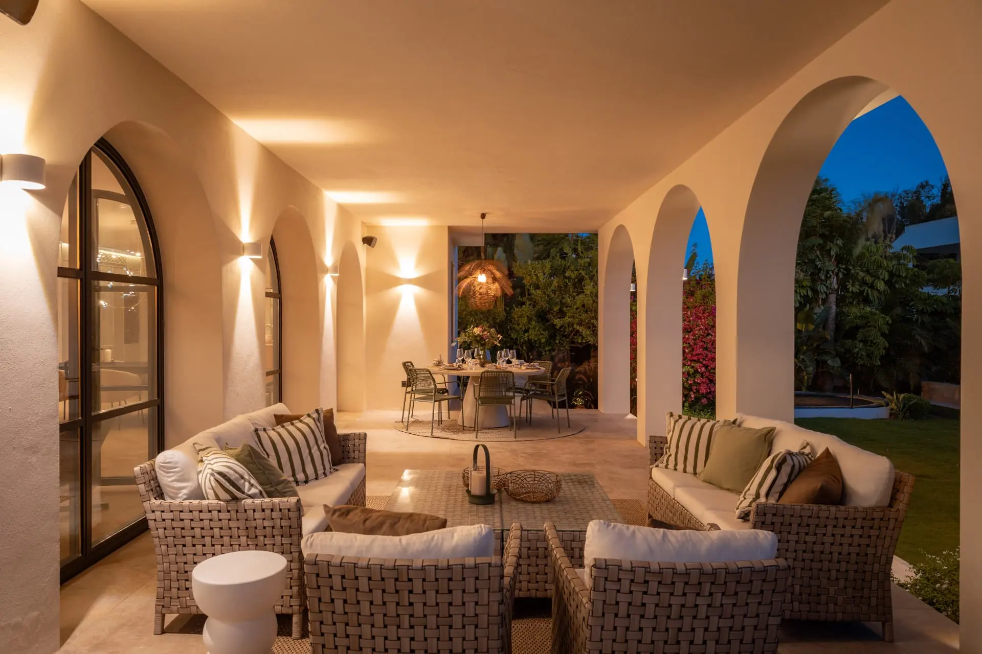The arched loggia at dusk, woven seating and a candlelit table by the garden
